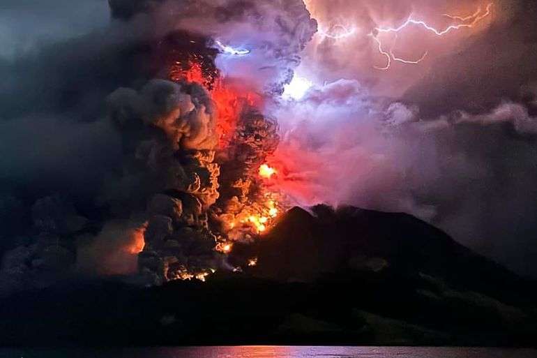 The Ruang volcano erupting. There are billowing clouds of smoke and ash obscuring the sun. Orange flames can also be seen.