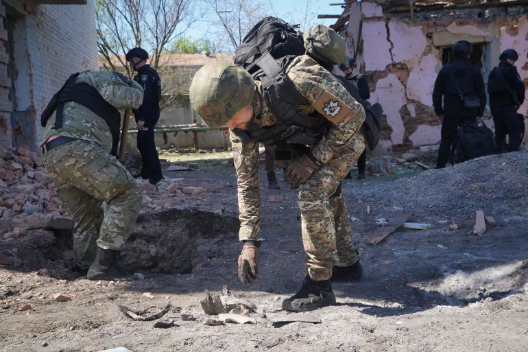 A policer officer bending down to pick up fragments from a Russian guided bomb that hit Kharkiv. Others behind him are also investigating the scene. There are piles of rubble and debris. A building behind has cracked walls and missing masonry.