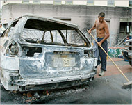 A man sweeps up broken glass near a burned out car