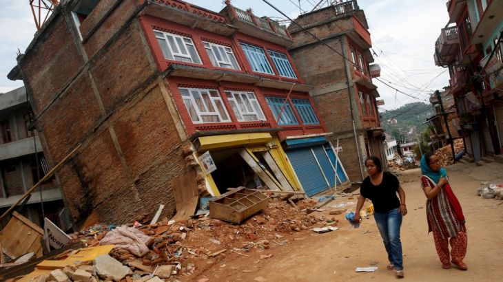 Women walk past a building damaged by earthquakes, causing it to lean to a side, in Sindhupalchowk district