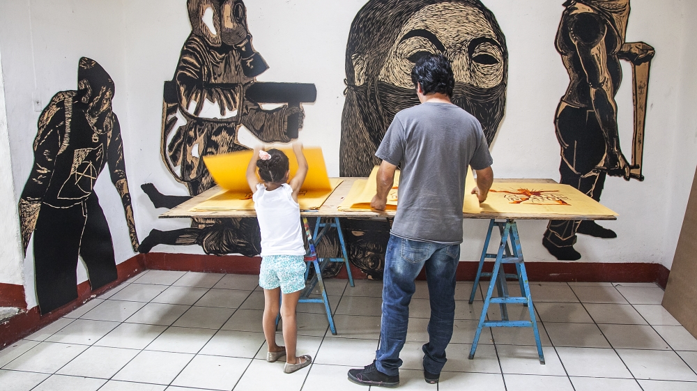 
Mario and his daughter count and sort screen prints for pasting on the city walls. Behind them, large wood prints depict images of protest [Gabriela Campos/Al Jazeera] 
