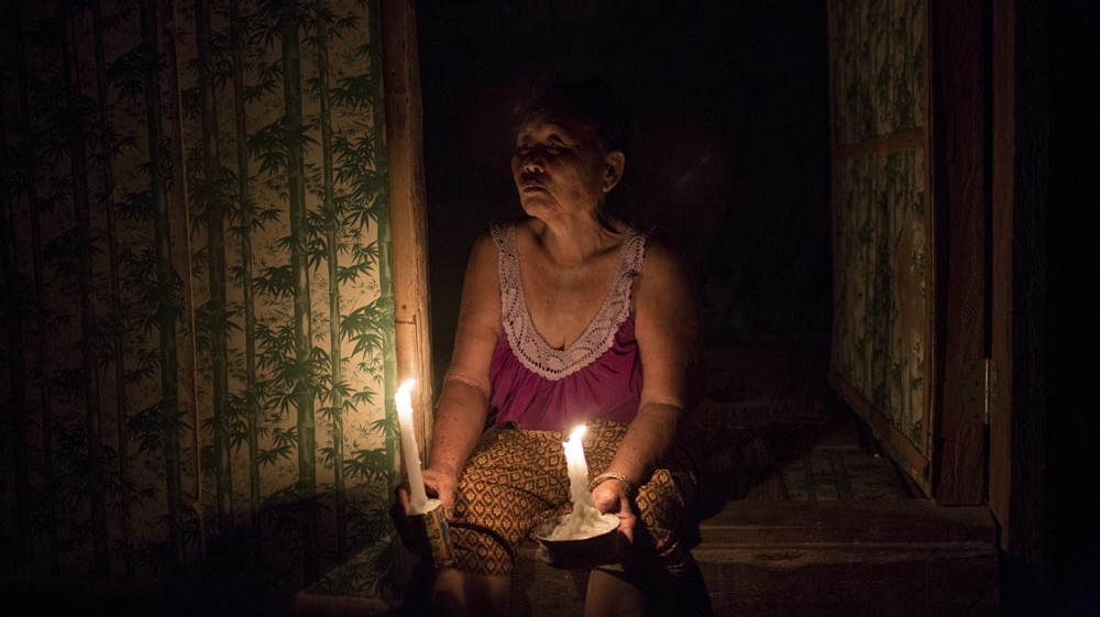 Rattana Seli, 66, sits at the entrance to her hut holding two candles. The village has no electricity so at night mostly candles are used [Luke Duggleby/Al Jazeera]
