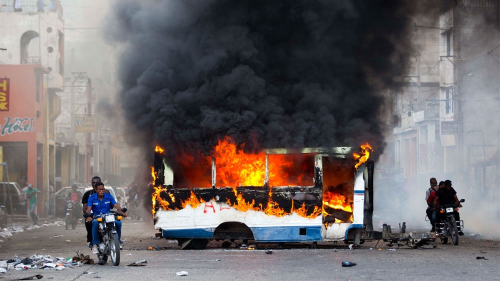 Motorcyclists pass by a burning bus, set on fire by opposition protesters in Port-au-Prince [Dieu Nalio Chery/AP]