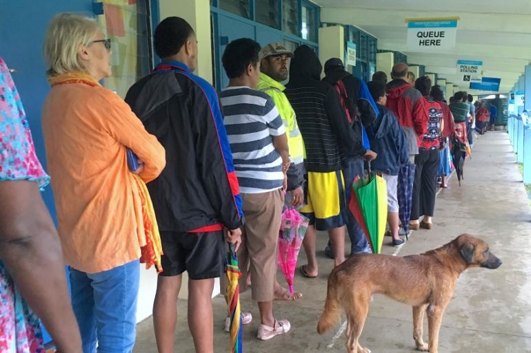 A dog stands with voters as they wait in a line to enter a polling venue near Suva