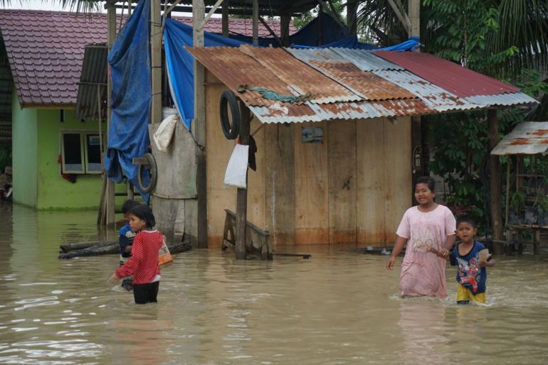 Children stand in floodwaters in front of a wooden building with a rusted corrugated roof in Aceh