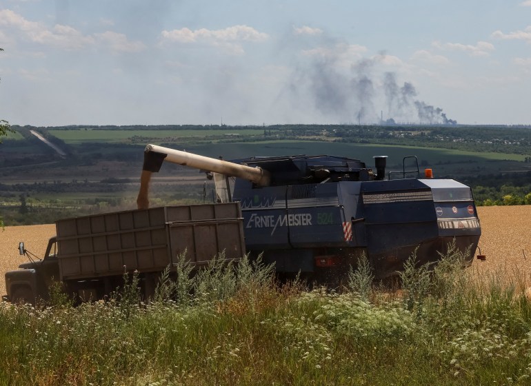 Farmers harvest wheat as Vuhlehirsk's heat power plant burns in the distance after a shelling, amid Russia's attack on Ukraine, in the Donbas region