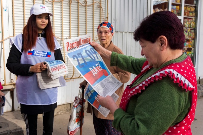 A woman looks at a newspaper ahead of the vote to join Russia that is due to start on Friday, watched by a young woman who is distributing the papers and an elderly woman