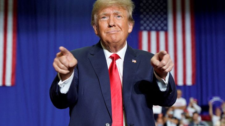 Donald Trump in suit and red tie, pointing with both hands with US flag behind him