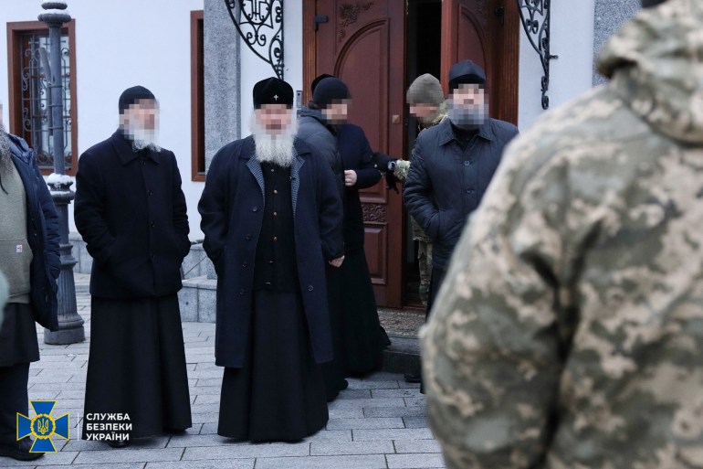 a grouo of Orthodox Christian priests in black robes and with long grey beards stand outside the Kyiv Pechersk Lavra monastery. Ukrainian intelligence service agents and police are also in the pictue, One wearing fatigues is standing with back to the camera in the foreground