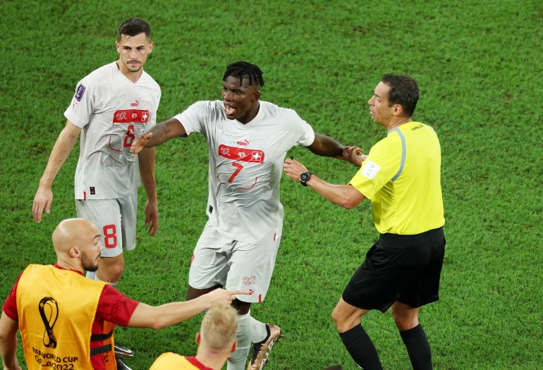 Embolo looking angry and gesturing at the referee who is pointing somewhere on the field