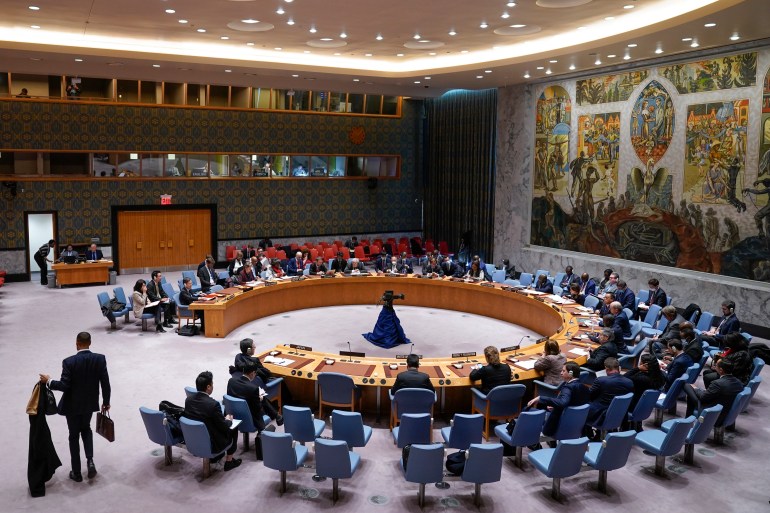 A wide view of the UN Security Council chamber. There are delegations around a central horseshoe shaped table and a large mural on the wall behind the table.