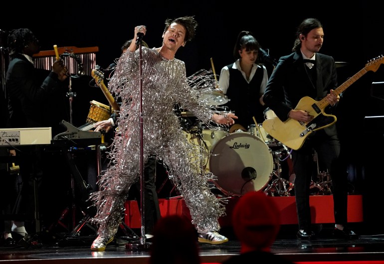 Harry Styles singing As it Was at the Grammys. He is wearing a jumpsuit of silver fringed lame and is dancing, There's a band behind him