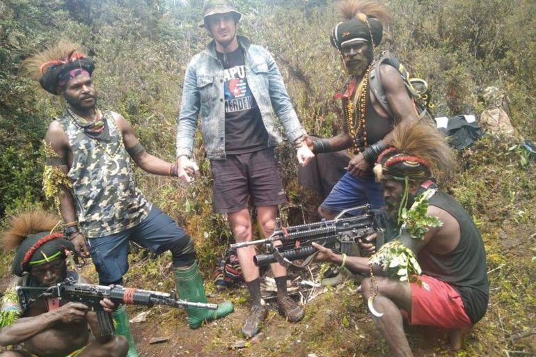 A man said to be Phillip Mehrtens surrounded by rebels in a rural location. He is wearing a t-shirt for Papuan independence, denim jacket, brown shorts and a hat. One rebel fighter is holding his wrist and another his hand. There are two fighters squatting at the front holding their guns.