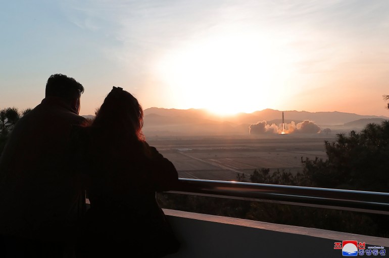 Kim Jong Un and his daughter watching the missile launch from a balcony. They have their backs to the camera and the sun is setting from behind a mountain.