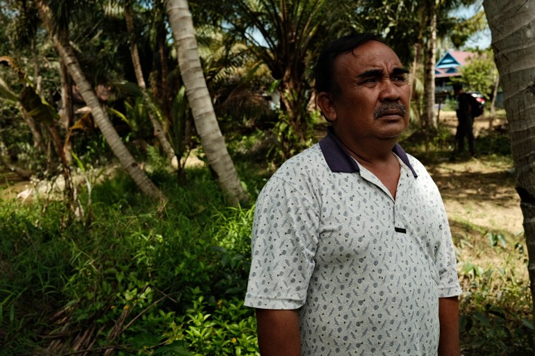 Atim standing in a grove of palm trees with houses in the distance. He is wearing a white polo short with a blue pattern and collar. He has a moustache.