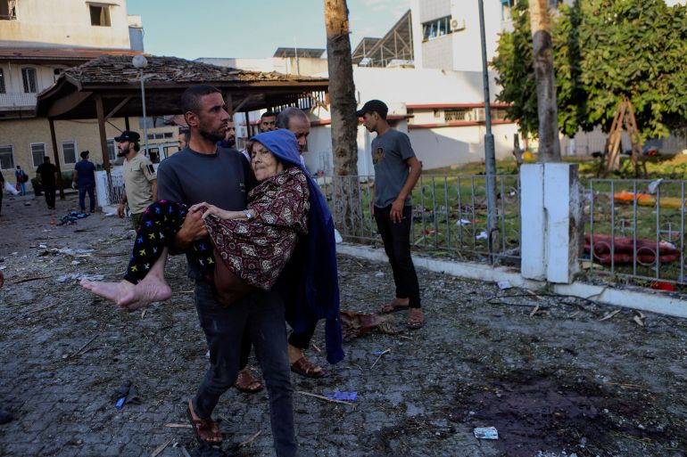 A Palestinian man carries an elderly woman past the site of a deadly explosion at al-Ahli hospital