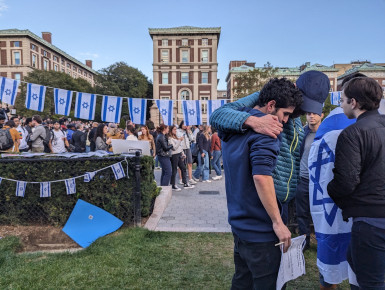 On a grassy lawn at Columbia University, a group of male students wrap their arms around each other's shoulders and hug, their heads bowed. One wears an Israeli flag. A line of Israeli flags can be seen behind them.