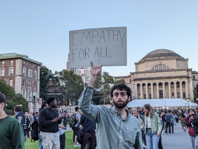 Standing in front of a domed building with columns in the front, a student protester holds up a sign written on cardboard that reads, "Empathy for all."