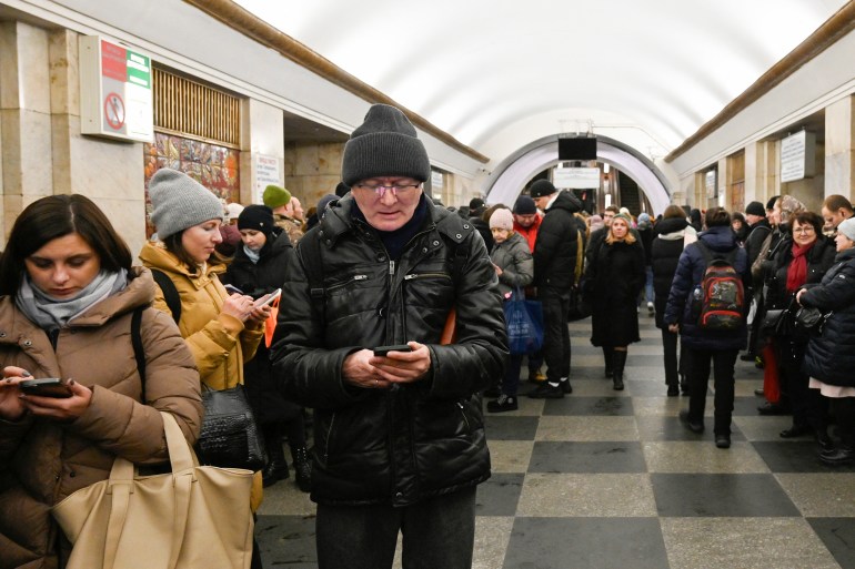 People taking shelter in the Kyiv metro during an air rad. They are dressed in winter clothes. Some are looking at that phones.
