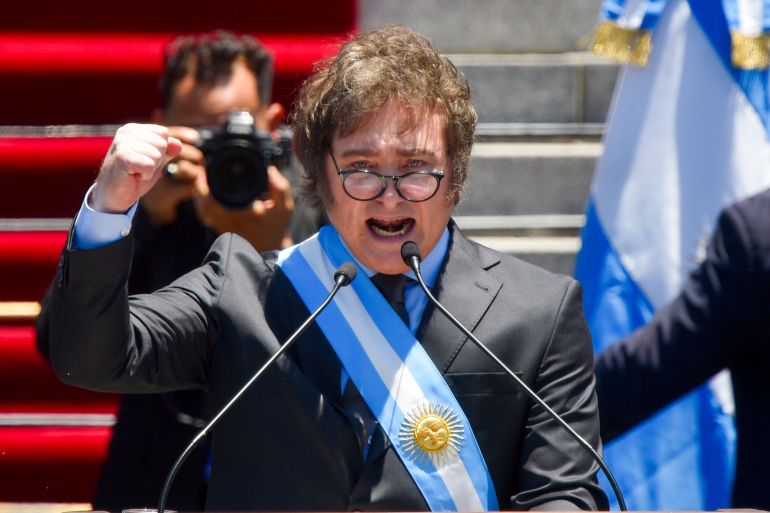 Javier Milei, shortly after being sworn in, speaks into the microphones at a podium outside Congress. He raises a fist as he speaks, wearing a dark suit, glasses and a sash in the colours of the Argentine flag. Behind him, a red carpet is visible on a set of granite steps.