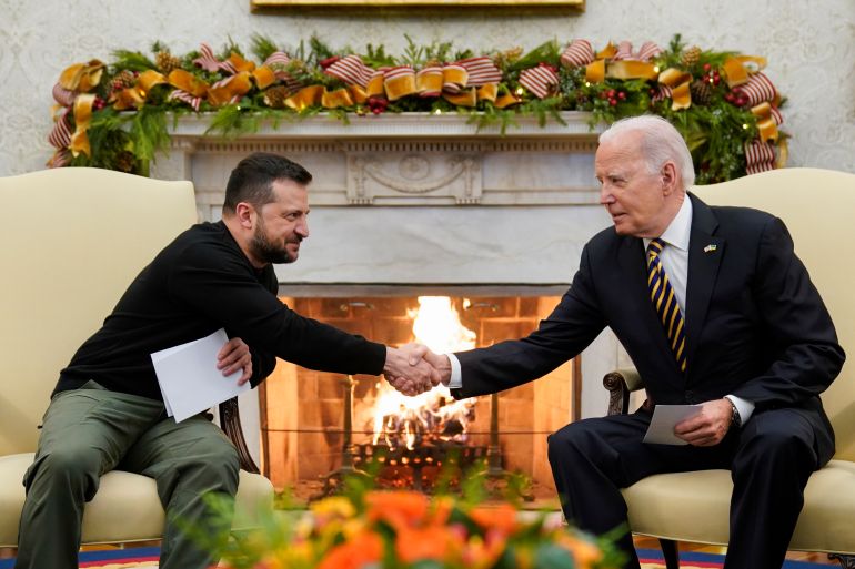 President Joe Biden and Ukrainian President Volodymyr Zelenskyy shake hands in the Oval Office. There is a fire behind them and a display of orange flowers and greenery across the mantlepiece. They are smiling.