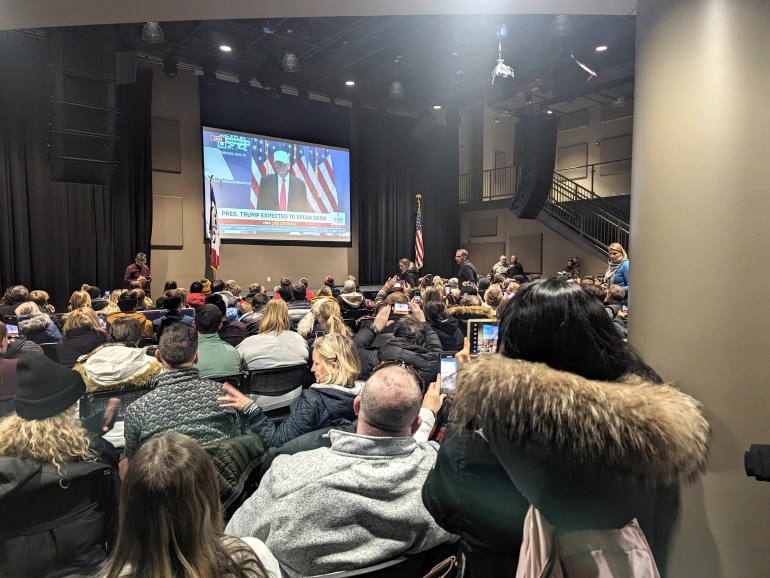 Trump supporters in a crammed room watch the former president speaking on a screen