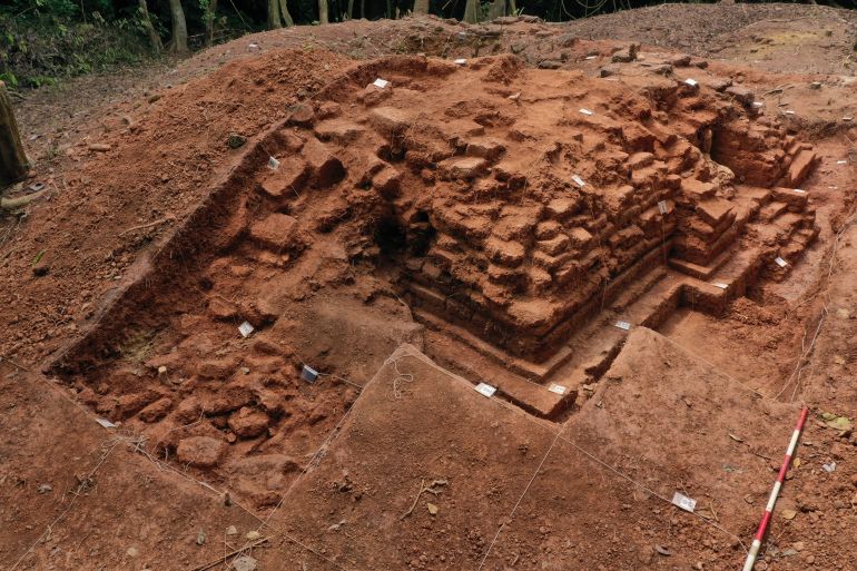 Excavation at Bukit Choras, A wall can seen emerging from the red earth. There are markers at various points. The jungle can be seen behind.