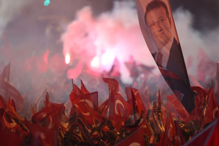 Crowd of people wave Turkish flags, with larger banner of Ekrem Imamoglu and red flares in the background