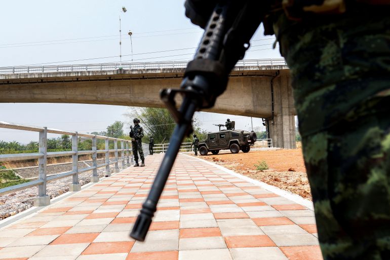 a view of the Thai Myanmar Friendship Bridge through a frame created by a soldier's rifle and his body. There is another soldier closer to the bridge as well as an armoured vehicle.