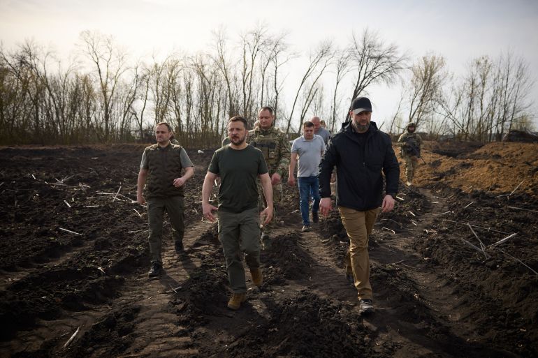 Ukrainian President Volodymyr Zelenskyy inspecting fortifications in the Kharkiv region. He s walking in mud with otjher officials. The landscape is brown and there are trees behind.