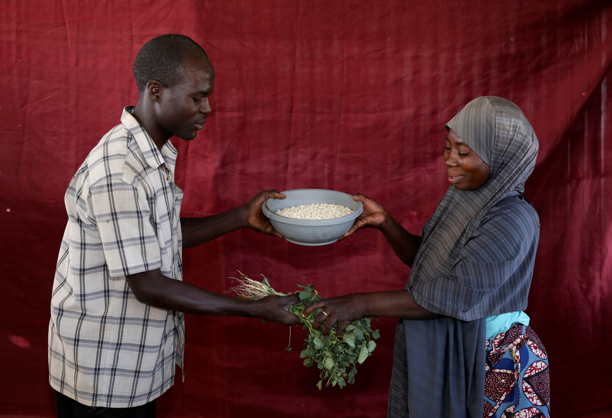 Falmata Ahmadu swaps her bowl of maize for Musa Ali Wala's amaranth vegetables. In Bakasi, the displaced frequently swap the little they have for their preferred goods, whether it's herbs and spices to make soup, groundnut to help a breastfeeding mother produce milk or laundry detergent for a family's clothes. [Afolabi Sotunde/Reuters]