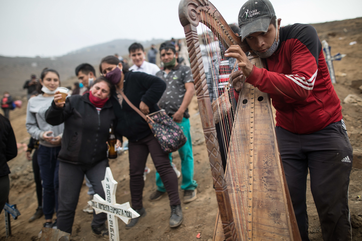Harpist and cemetery worker Charlie Juarez plays music as Gregoria Zumaeta, left, mourns the death of her brothers Jorge Zumaeta, 50, and Miguel Zumaeta, 54, who died of COVID-19. [Rodrigo Abd/AP Photo]