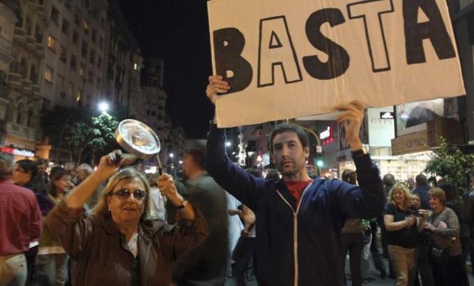 A protester bangs a pan as another holds a placard that reads