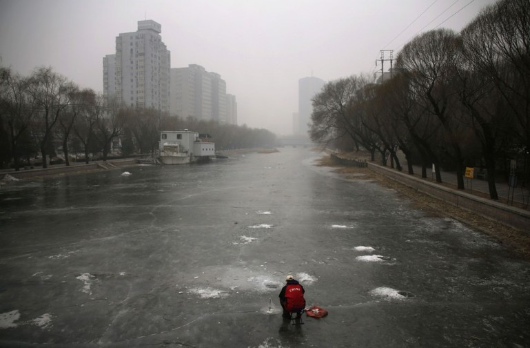 A man sits on a frozen canal and fishes on a hazy winter day in central Beijing