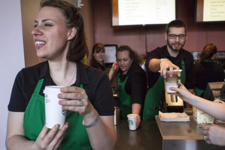 Barista in green apron holding coffee and smiling while behind her, other baristas hand coffees over the counter to customers