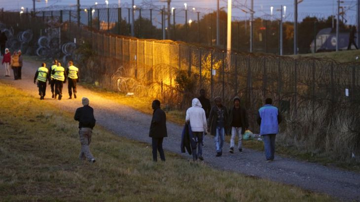 Migrants walk past a fence as another one tries to go in Eurotunnel freight shuttle near the Channel Tunnel access in Coquelles, near Calais