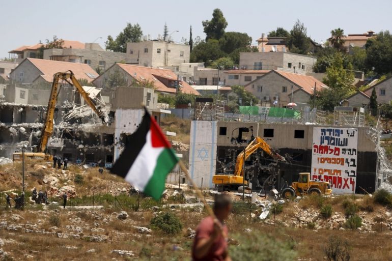 Palestinian man holds a Palestinian flag as Israeli heavy machinery demolish vacant apartment blocs by order of Israel''s high court, in the West Bank Jewish settlement of Beit El near Ramallah