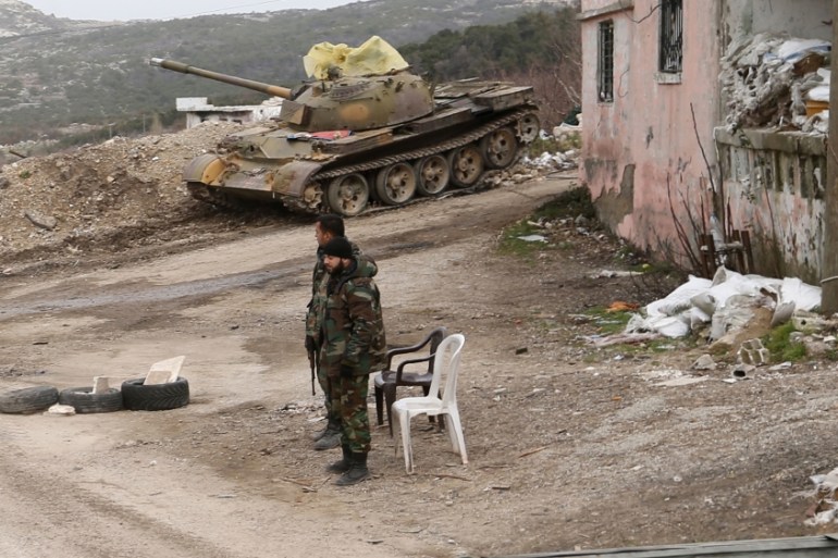 Syrian troops stand with a destroyed tank in the back