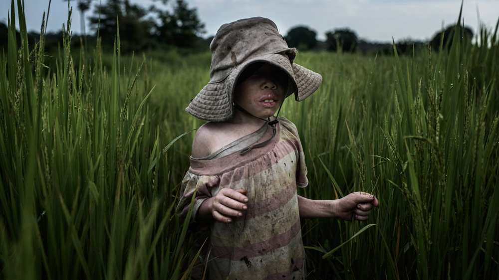 Semabulia plays in the rice field where the kids spend most of their time. His wide-rimmed hat protects his skin from the sun during the day [Fredrik Lerneryd/Al Jazeera]
