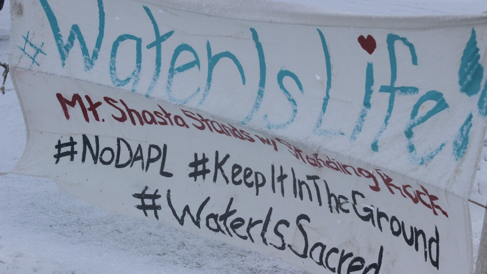 A banner along Flag Row echoes the camp's greeting-cum-battle cry, Mni Wiconi! - water is life. Hashtags connect the Lakota phrase to the social media bloodstream [Adam Levinson/Al Jazeera]