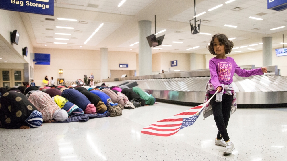 A young girl dances with an American flag in baggage reclaim while women pray behind her during a protest against the travel ban at an airport in Dallas, Texas [Reuters]