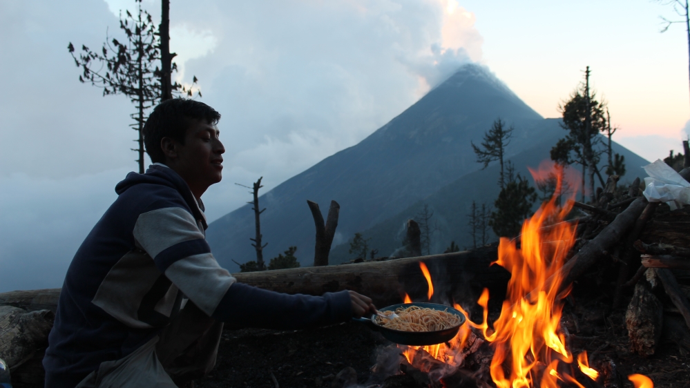 Elvin Soy Lopez, Aprode guide, prepares spaghetti after setting up camp on the Acatenango Volcano. The Volcano of Fire erupts in the background [Martha Pskowski/Al Jazeera]