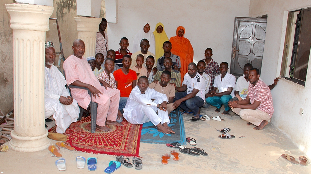 Alhaji Ibrahim Abbas (seated on the chair), his first wife Bilkisu (in the orange hijab) and his other wives are photographed with the people they are currently hosting. Butcher Umar Guwa (who wears a white football jersey) used to live there and David John, to his left in the shirt in the green stripes, currently stays with Abbas' family [Femke van Zeijl/Al Jazeera]
