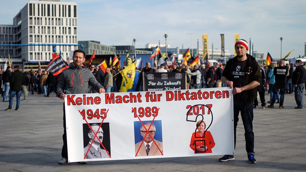 Thomas Witte, right, holds up a banner likening German Chancellor Merkel to Adolf Hitler and the GDR's Erich Honecker [Sorin Furcoi/Al Jazeera]