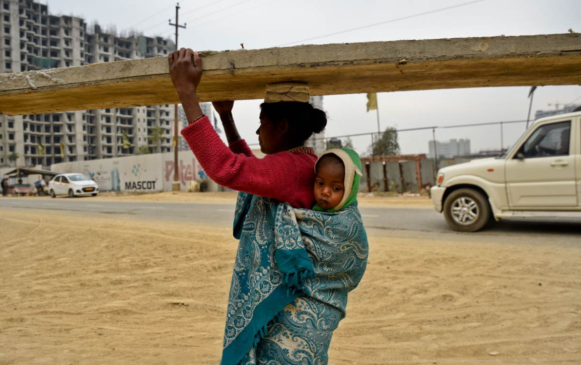 An Indian woman working at a building construction site has her eighteen month child wrapped on to her back as she walks carrying a heavy construction material on her head in Greater Noida, near New D