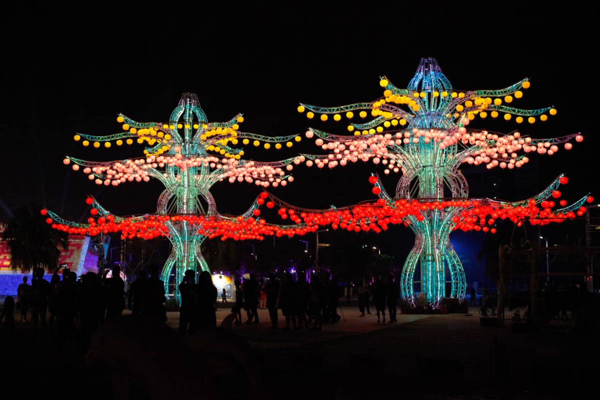 People walk under several traditional ball-shaped lanterns hanging on the intertwined branches of trees lit with modern lighting and design. Inspired by hand-dyed Indian tapestry art from the National