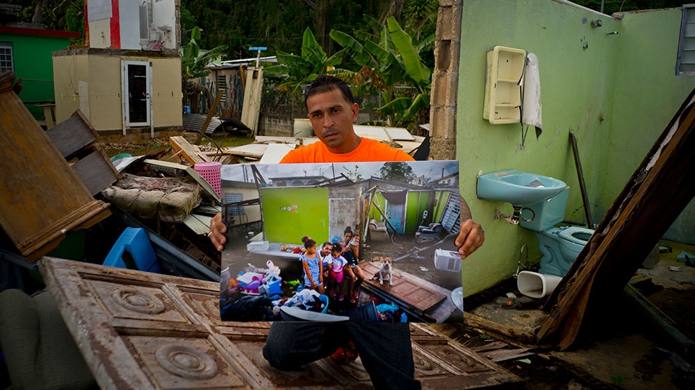 Arden Dragoni holds a photo taken on October 5, 2017, of his home that was destroyed by Hurricane Maria, as he stands in the same spot where his home remains in shambles in Toa Baja, Puerto Rico on May 28, 2018 [Ramon Espinosa/AP Photo]