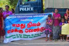 Protesters stand in front of a police truck that has been parked across the road from the gate, with a banner that reads "We want our land back" in Tamil [Lisa Fuller/Al Jazeera]