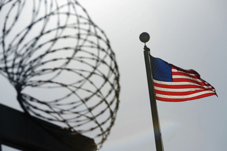 Barbed wire with the US flag to the right of it. The wire and fence is black, like a silhouette against the sky.