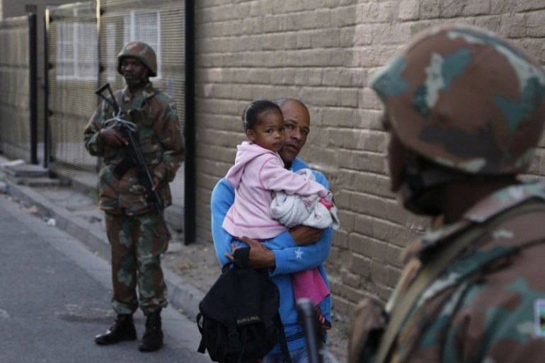 A man holding a girl walks by two South African soldiers providing security for police officers during a raid in Manenberg, South Africa Thursday, May 21, 2015